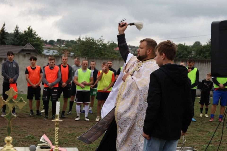 Священик кропить святою водою поле, поруч хлопець і футболісти